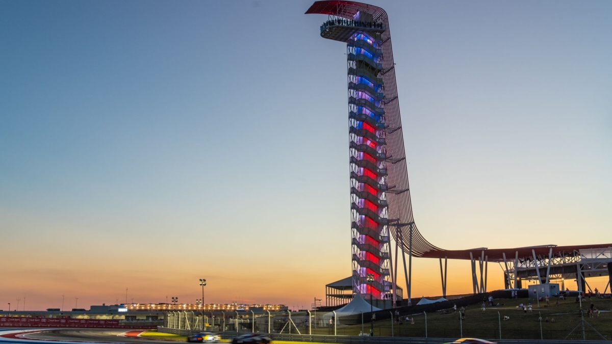 مسابقه فرمول یک در پیست COTA آستین تگزاس با برج قرمز نمادین در پس‌زمینه؛ خودروهای مسابقه‌ای در حال عبور از پیچ با جدول‌های رنگی قرمز، سفید و آبی، نور غروب خورشید و حضور تماشاگران در فضای باز، نماد هیجان موتوراسپرت آمریکا.